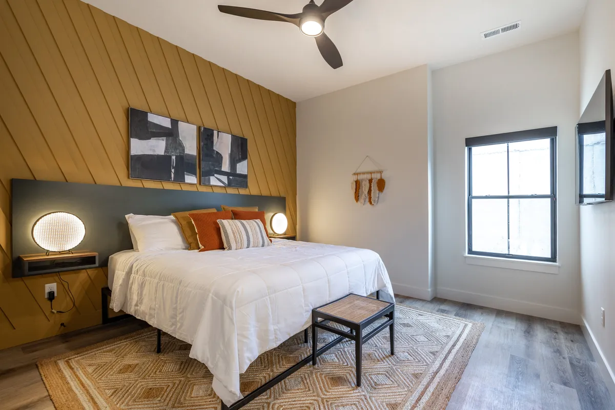 A tranquil bedroom adorned with white bedding and decorative pillows, geometric wall art, sophisticated nightstand lamps, and a wooden bench at the foot of the bed. Natural light streams in from a large window, creating a restful and inviting atmosphere.