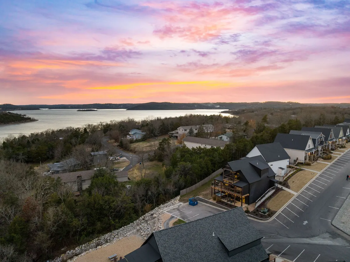 A tranquil view of a sunset sky over a water surface, with homes along the shore and natural landscape in the background. The serene ambiance invites relaxation, lakeside strolls, and appreciation of the changing seasons.
