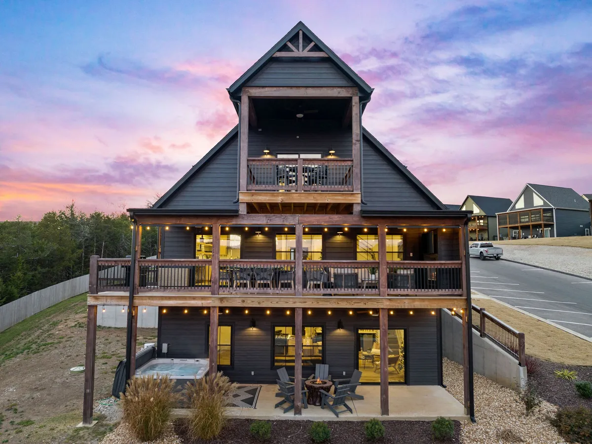 A wide exterior shot showcasing the multi-level deck with a hot tub, comfortable outdoor seating, and the home's distinctive triangular roof. The deck overlooks scenic views, with sunset lighting creating a welcoming and cozy ambiance.
