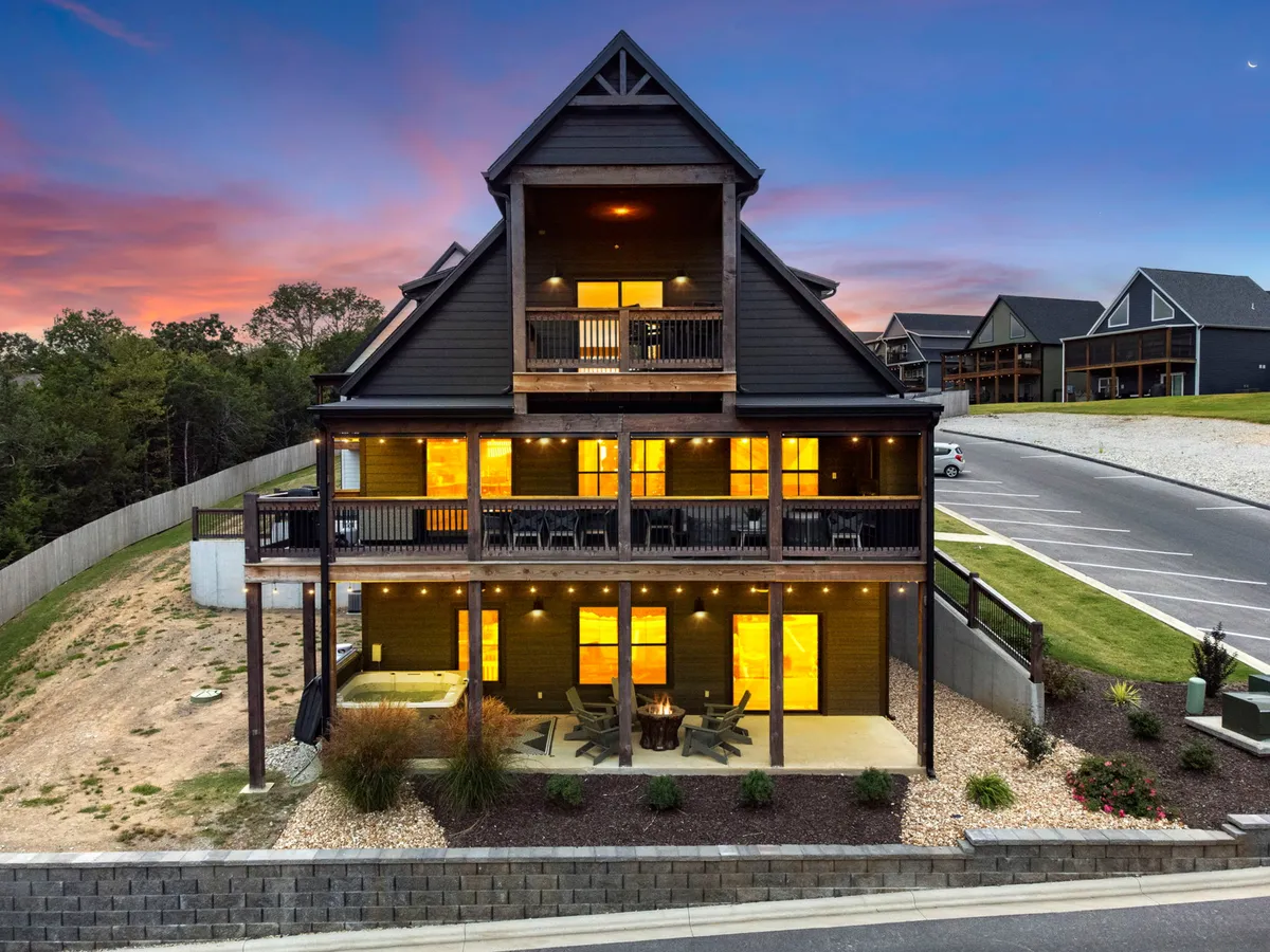 A warmly illuminated exterior view of Keystone Lodge at dusk. The large deck is set against a stunning mountain backdrop, with landscaped gardens visible. The scene features areas for outdoor dining and space for gatherings, all glowing with a welcoming light.