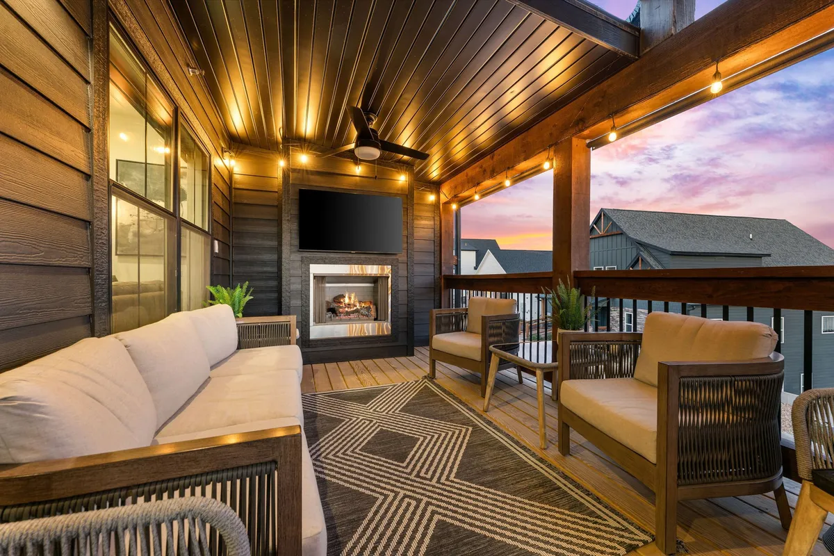 The image showcases a stylish outdoor living area with a warm fireplace, plush lounge seating arranged on a chic decorative rug, a mounted TV for entertainment, all set under the glow of string lights. The inviting setup overlooks serene lake views, perfect for unwinding or celebrating together as the sun sets.