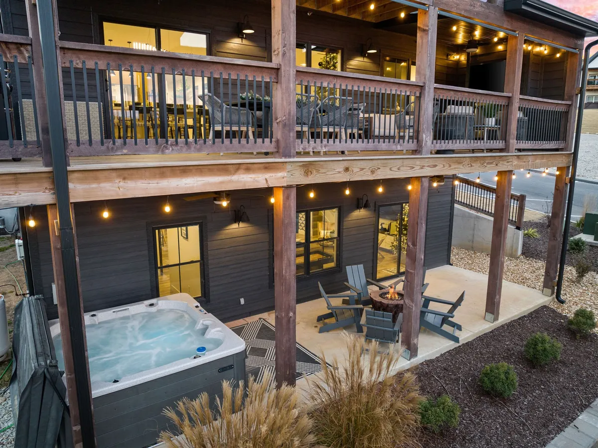 A cozy outdoor deck featuring a hot tub, comfortable seating area, string lights, and wooden decking. The inviting ambiance is perfect for evening relaxation, gatherings by the fire pit, and stargazing from the balcony.