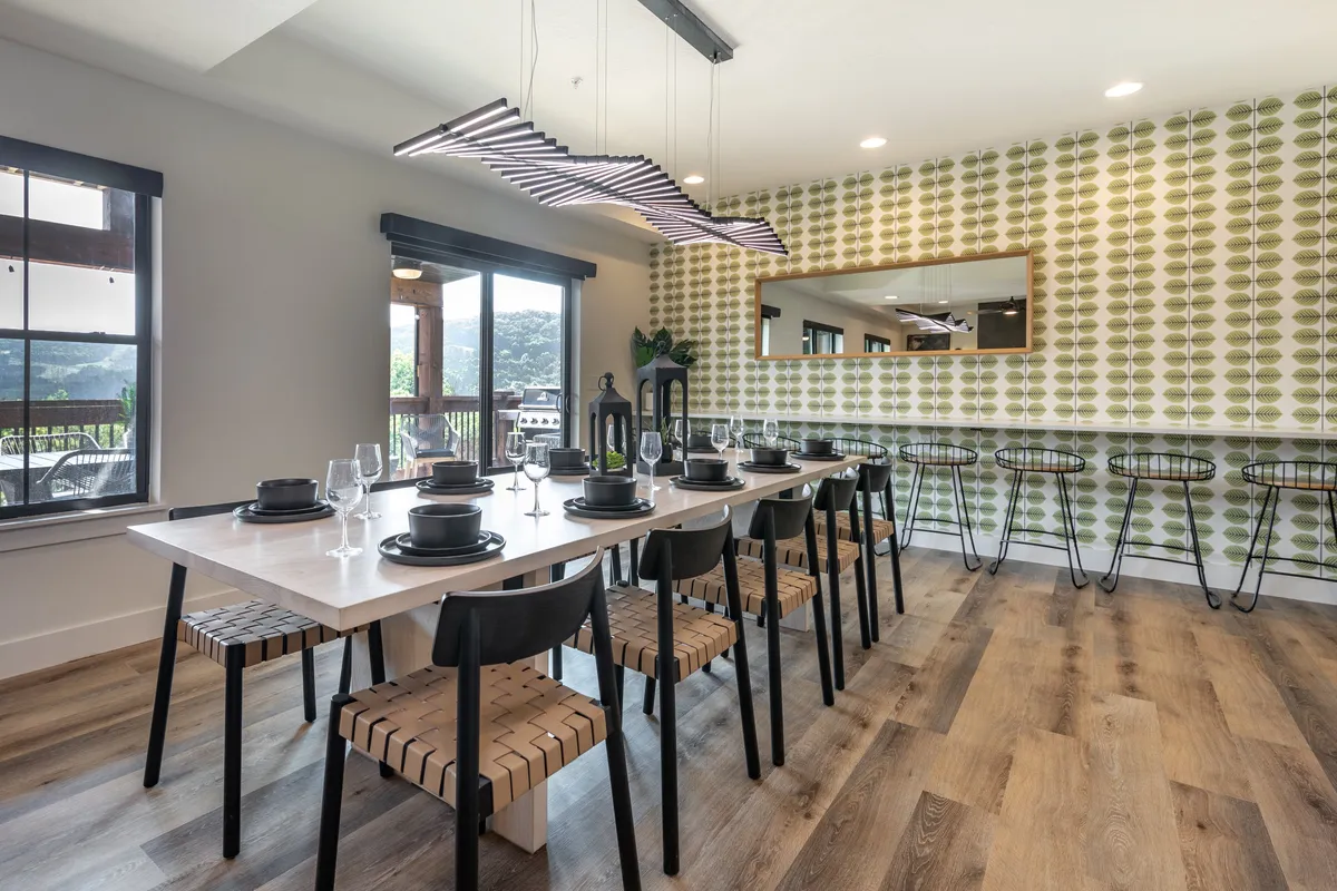 This photo captures a long dining table set beneath a unique hanging light fixture, with a sophisticated green patterned wall and a reflective mirror in the background. The table is ready for large family dinners or festive celebrations.